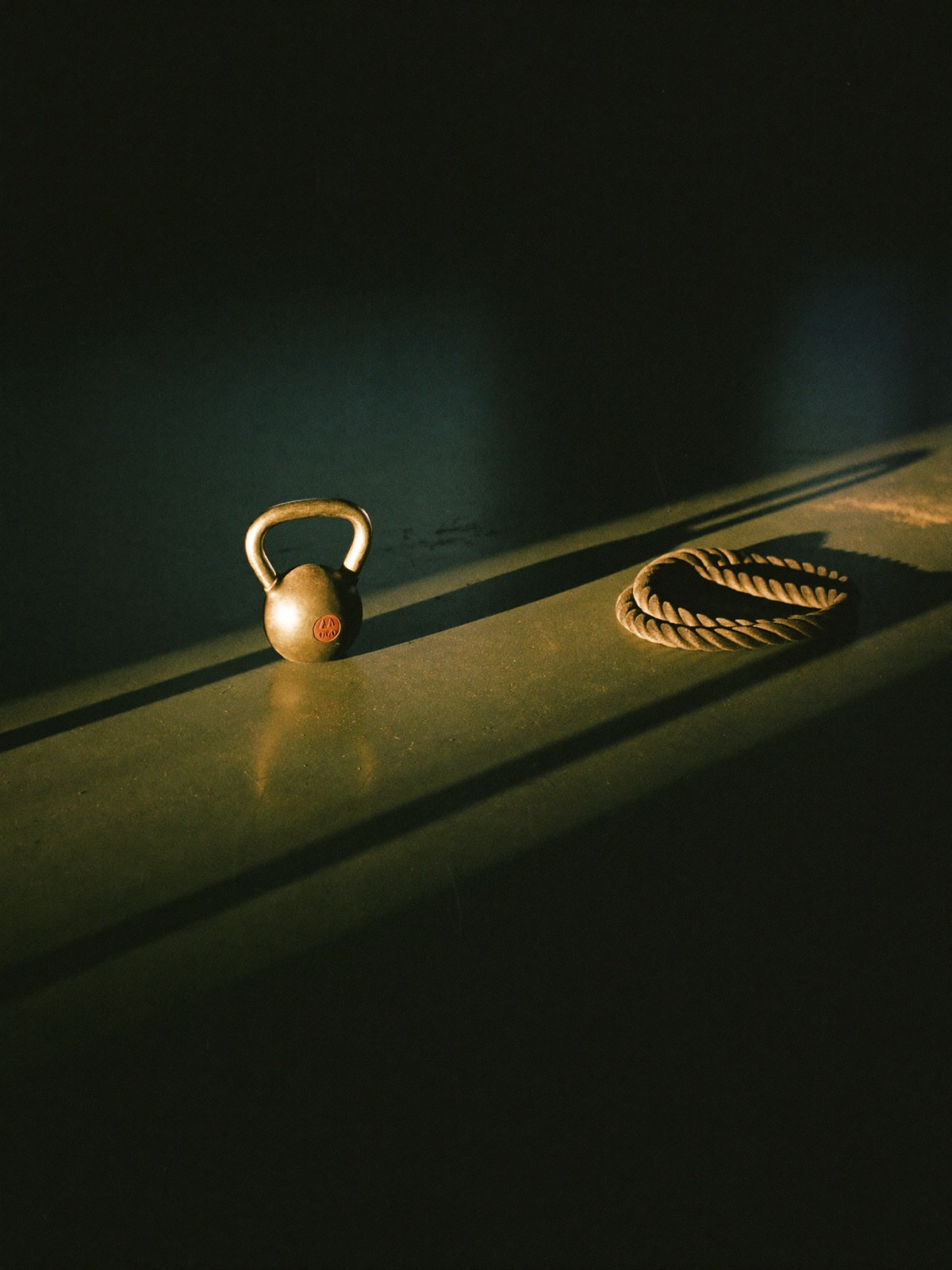 A cast-iron kettlebell and coiled rope in a dark gym, crossed by one shaft of golden light