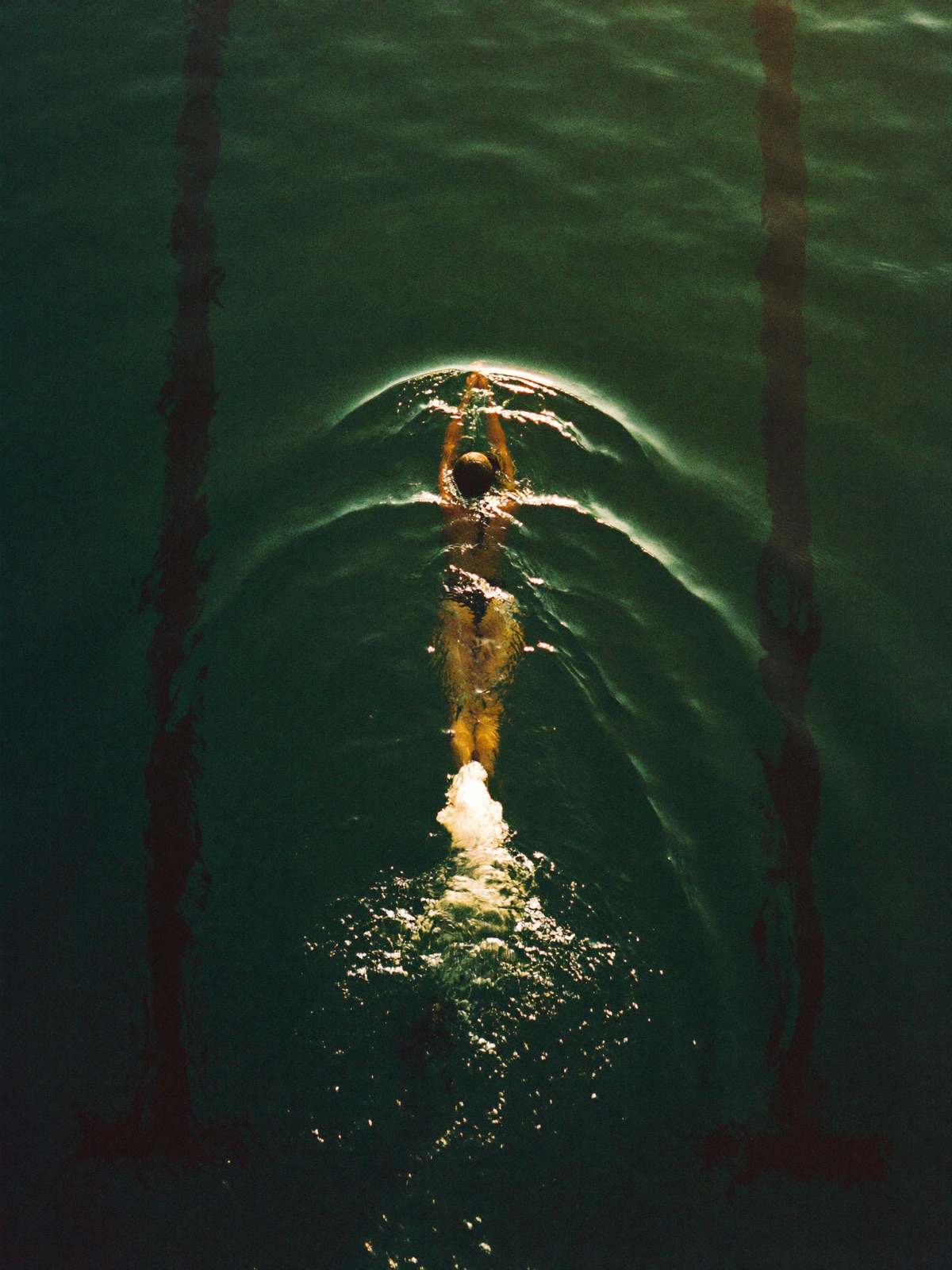 A single swimmer seen from above in a dark emerald pool crossed by golden morning light