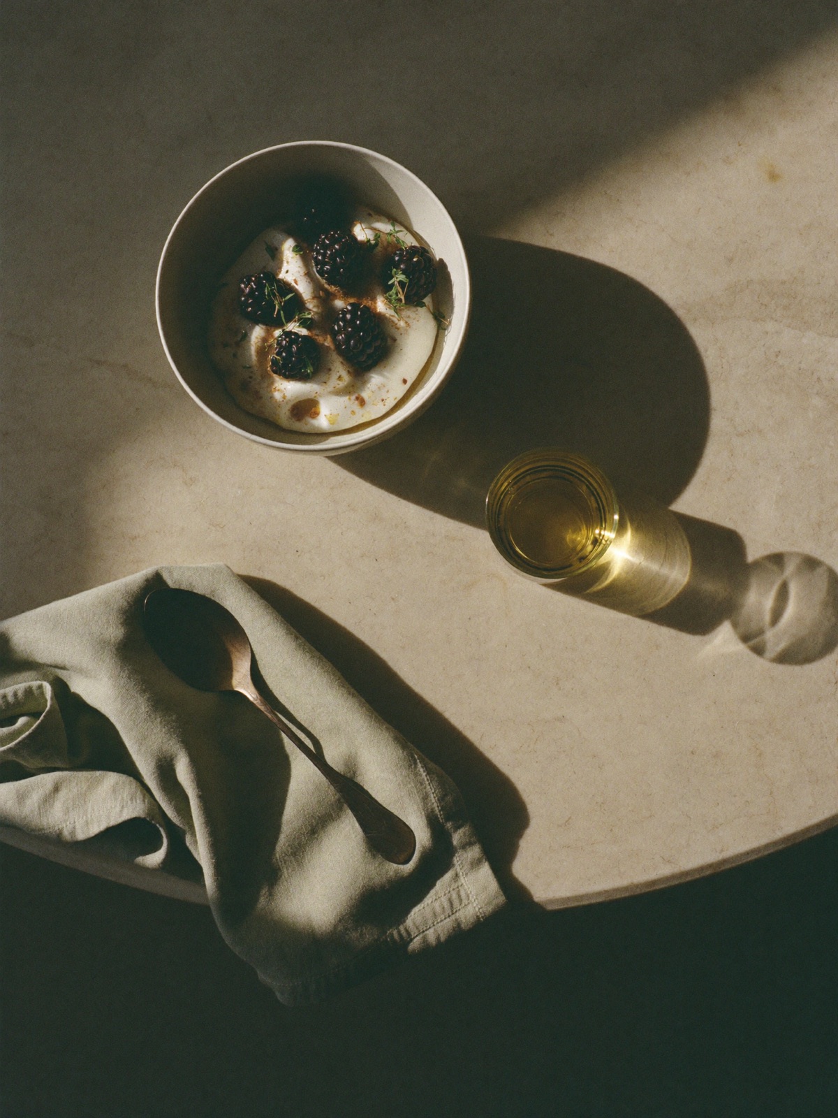 Overhead view of a minimal breakfast — yogurt with berries, olive oil and a sage linen napkin on warm stone